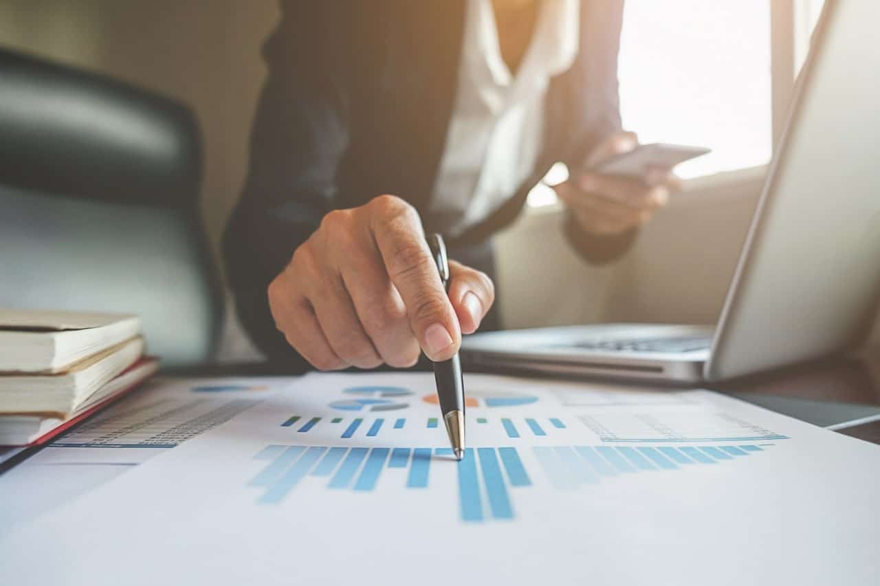 Das Bild zeigt eine Person in einer Geschäftsumgebung, die mit einem Stift auf ein Dokument zeigt. Das Dokument enthält verschiedene Diagramme und Grafiken, die sich möglicherweise auf Finanz- oder Geschäftsdaten beziehen. In der anderen Hand hält die Person auch ein Smartphone, auf dem Schreibtisch liegen ein Laptop und mehrere Bücher.