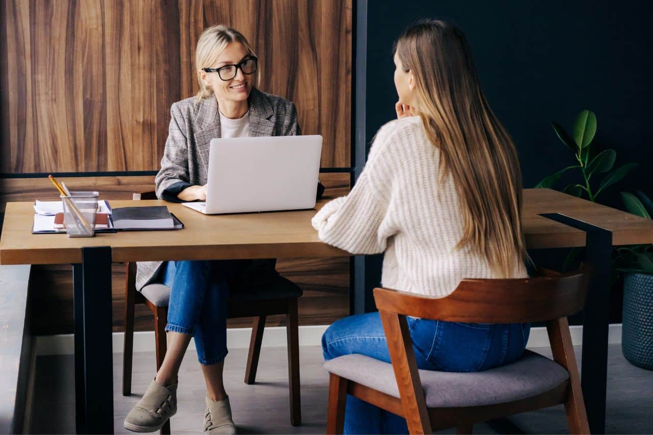 Das Bild zeigt ein professionelles Gespräch zwischen zwei Frauen in einem modernen Büro. Eine Frau mit blonden Haaren und Brille sitzt an einem Schreibtisch und trägt ein kariertes Sakko. Sie schaut freundlich auf die andere Frau, die mit dem Rücken zur Kamera sitzt und einen weißen Strickpullover trägt. Auf dem Tisch befinden sich ein aufgeklappter Laptop, ein Notizbuch, Schreibutensilien und ein Glas. Im Hintergrund ist eine schlichte, elegante Holzwand und eine Pflanze zu sehen. Die Szene vermittelt eine professionelle und angenehme Atmosphäre, möglicherweise in einem Bewerbungsgespräch.