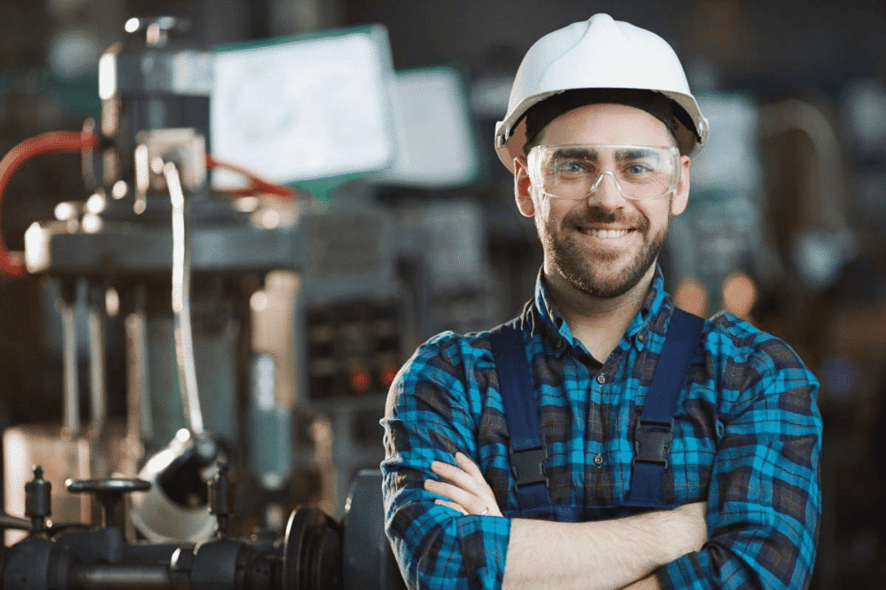 Das Foto zeigt einen Mann, der in einer industriellen Umgebung arbeitet. Der Mann trägt einen weißen Schutzhelm, ein blaues kariertes Hemd mit Hosenträgern und verschränkte Arme. Im Hintergrund sind verschiedene Industriemaschinen und -anlagen zu sehen.