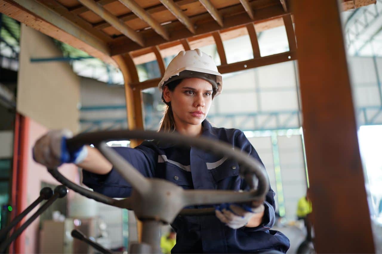 Das Bild zeigt eine Frau in Arbeitskleidung und einem Schutzhelm, die ein Fahrzeug, vermutlich einen Gabelstapler, steuert. Sie konzentriert sich auf ihre Aufgabe und befindet sich in einer industriellen Umgebung, wie einer Lagerhalle oder Fabrik. Die Szene vermittelt Professionalität und Engagement in einem Arbeitsumfeld.