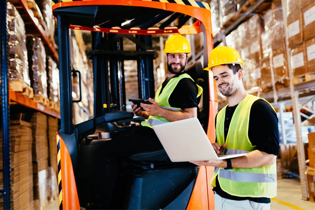 Auf dem Bild sehen wir zwei lächelnde Mitarbeiter im Lager, beide mit gelben Helmen und Warnwesten gekleidet. Einer der Mitarbeiter sitzt in einem Gabelstapler und hält das Gerät in der Hand, während der andere vor ihm steht und einen Laptop bedient. Die beiden scheinen eine Aufgabe zu besprechen oder Daten in einer Arbeitsumgebung mit Regalen voller Kisten zu überprüfen.