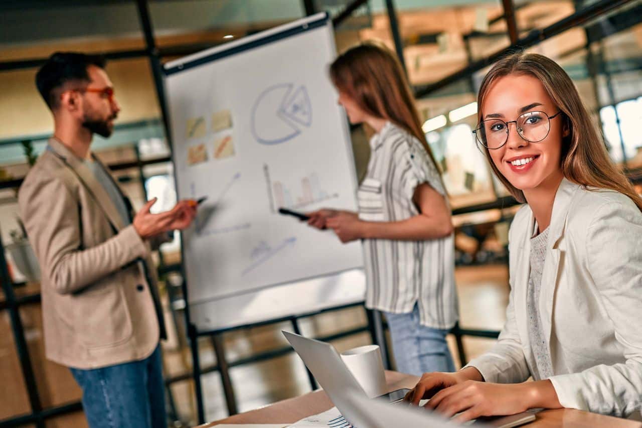 Das Gemälde zeigt drei Menschen in einem modernen Büro. Eine von ihnen ist eine Frau, die an einem Schreibtisch sitzt und an einem Laptop arbeitet, und die anderen beiden - ein Mann und eine Frau - stehen neben einem Whiteboard. Auf dem Board sehen Sie verschiedene Diagramme und Grafiken, darunter Kreisdiagramme, Balkendiagramme und Liniendiagramme. Die Frau, die am nächsten an der Tafel steht, zeigt mit einem Marker darauf und erklärt oder präsentiert die Informationen wahrscheinlich den anderen.
