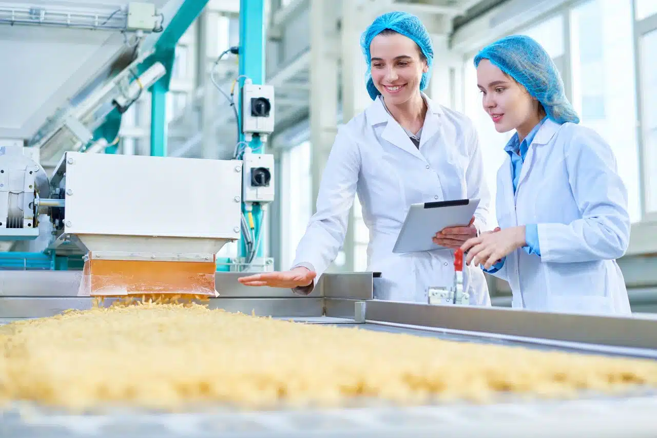 Das Gemälde zeigt zwei lächelnde Frauen in einer Fabrik. Sie tragen weiße Schürzen und blaue Haarnetze, was die Bedeutung von Hygiene und Sicherheit in dieser Umgebung unterstreicht. Eine der Frauen hält ein Klemmbrett in der Hand, was darauf hindeutet, dass sie den Produktionsprozess überwacht oder kontrolliert. Im Hintergrund sind Maschinen zu sehen, die an der Herstellung von Lebensmitteln beteiligt sind, möglicherweise Nudeln oder anderen Lebensmitteln, die auf dem Förderband zu sehen sind. Dieses Bild unterstreicht die Bedeutung von Qualitätskontrolle und Sicherheit in der Lebensmittelindustrie
