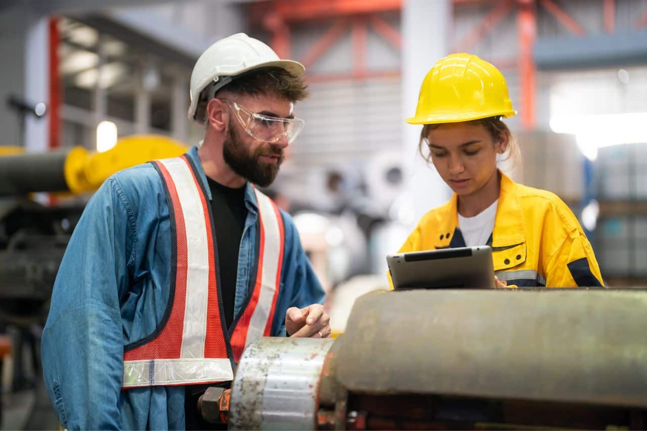 Das Gemälde zeigt einen Mann und eine Frau in einem Arbeitsumfeld, möglicherweise in der Industrie oder auf dem Bau. Beide tragen Schutzausrüstung, darunter Helme und Warnwesten. Eine Frau hält ein Tablet in der Hand, auf dem sie wahrscheinlich arbeitsbezogene Informationen diskutiert oder überprüft. Im Hintergrund sind Maschinen und andere Industrieanlagen zu sehen, was darauf hindeutet, dass es sich um einen Ort handelt, der eine technische Herangehensweise an die Arbeit erfordert. Dieses Bild unterstreicht die Bedeutung von Sicherheit und Zusammenarbeit in Industrie- oder Bauumgebungen.