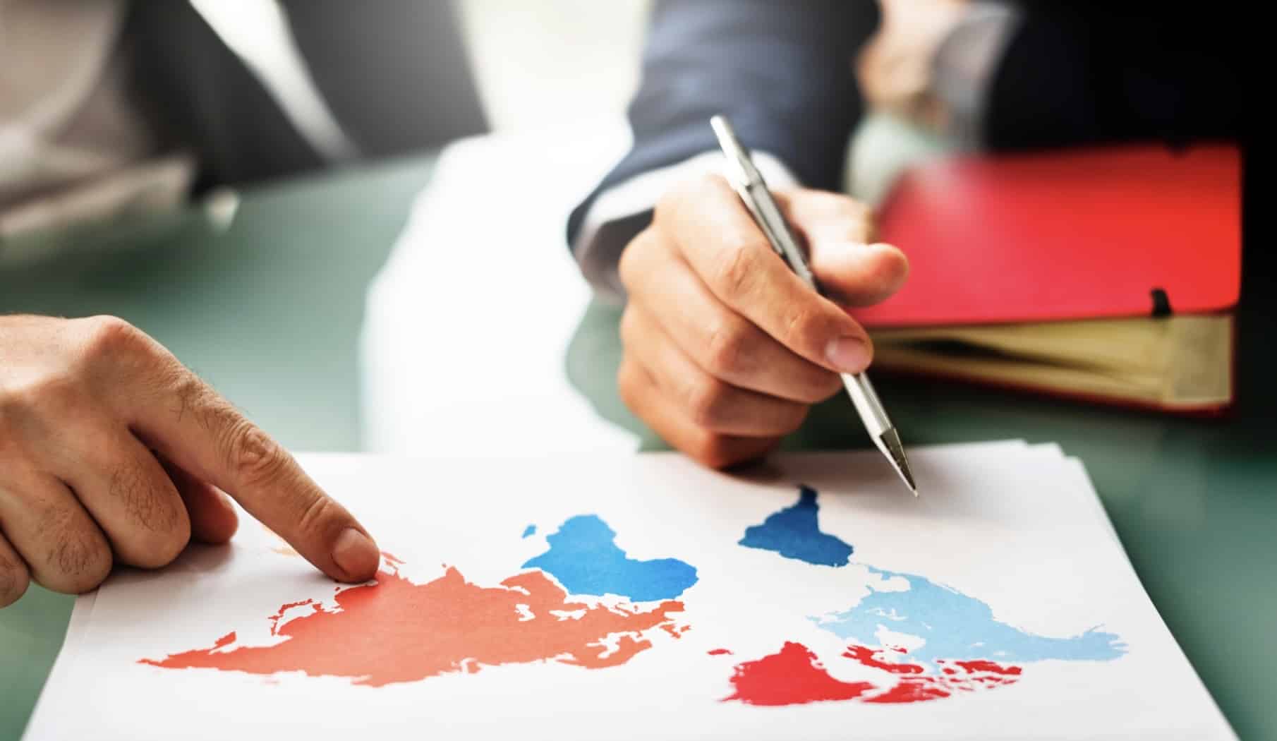 Auf dem Foto sieht man einen Ausschnitt eines Geschäftstreffens. Im Vordergrund sind zwei Hände zu sehen – eine Person zeigt mit dem Finger auf eine Weltkarte, während die andere einen Stift hält, der ebenfalls auf die Karte gerichtet ist. Die Karte ist auf ein Blatt Papier gedruckt und zeigt die Kontinente in den Farben Rot und Blau. Im Hintergrund ist ein Tisch und ein Notizbuch zu sehen, was darauf hindeutet, dass es sich um ein Gespräch über Strategien, Planung oder internationale Aktivitäten handelt.