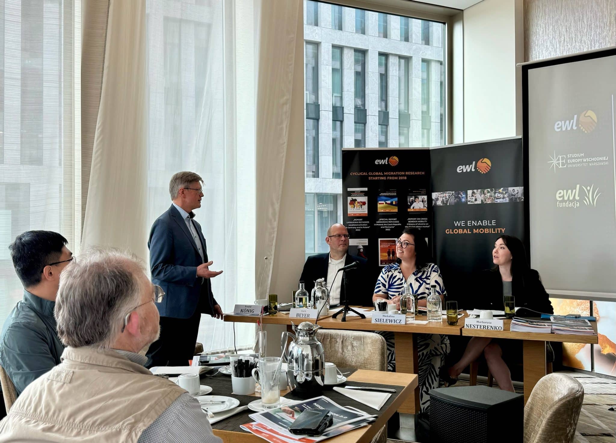 Das Bild zeigt ein Geschäftstreffen oder eine Konferenz, die in einem modernen Büro stattfindet. Mehrere Personen sitzen an einem Tisch und eine Person steht auf und hält eine Präsentation. Auf dem Tisch stehen Teller mit den Namen der Teilnehmer, im Hintergrund sind Banner und ein Bildschirm zu sehen, auf dem das Logo und der Slogan "ewl" zu sehen sind. Auf den Bannern stand "WE ENABLE GLOBAL MOBILITY" sowie verschiedene Bilder und Texte, die sich auf die Organisation beziehen. Die Umgebung ist professionell, mit Dokumenten, Gläsern und einem Mikrofon auf dem Tisch, was auf eine formelle Diskussion oder Präsentation hinweist.