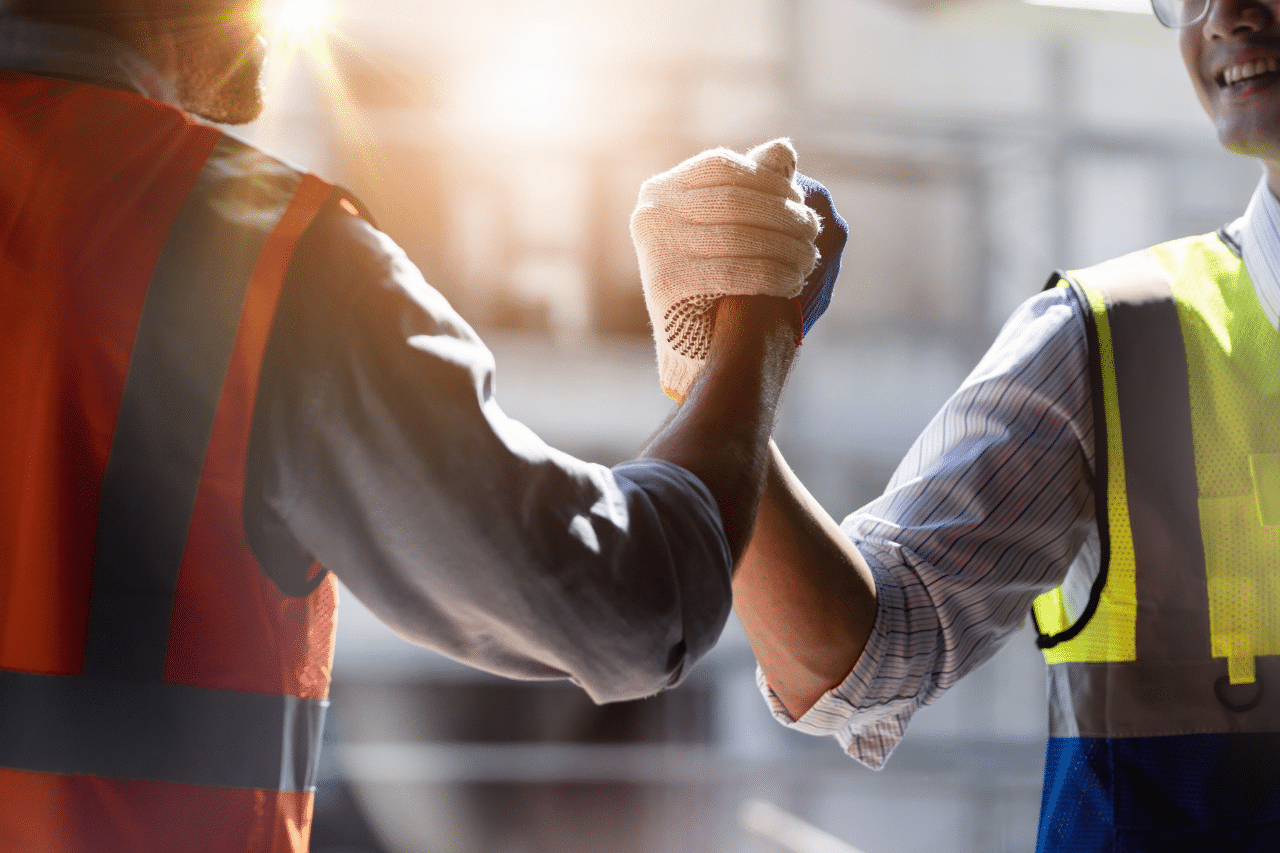Auf dem Bild sind zwei Personen in reflektierenden Westen und Arbeitshandschuhen zu sehen, die sich die Hände schütteln. Der Hintergrund ist unscharf, aber es scheint sich um eine Baustelle oder ein industrielles Umfeld zu handeln, was durch die Kleidung und die Umgebung angedeutet wird.