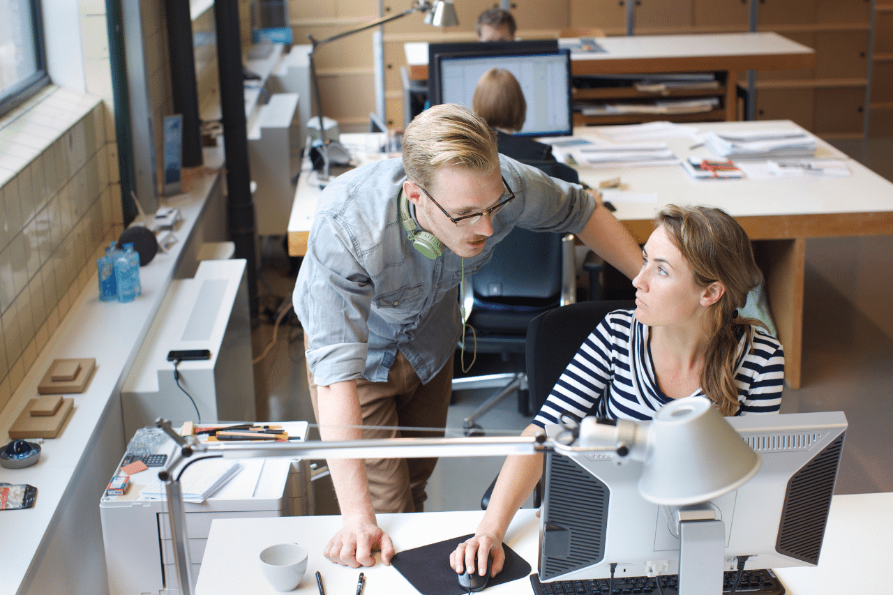 Auf dem Foto sind zwei Mitarbeiter im Büro zu sehen. Der Mann mit der Brille und dem hochgekrempelten Hemd beugt sich über den Tisch der Frau und weist auf etwas auf dem Computerbildschirm hin. Um seinen Hals trägt er grüne Kopfhörer. Die Frau, die am Tisch in einem schwarz-weißen gestreiften T-Shirt sitzt, schaut ihn an, und ihre Hand liegt auf der Maus. Beide sind bei der Arbeit und es sieht so aus, als würden sie über ein Projekt diskutieren.