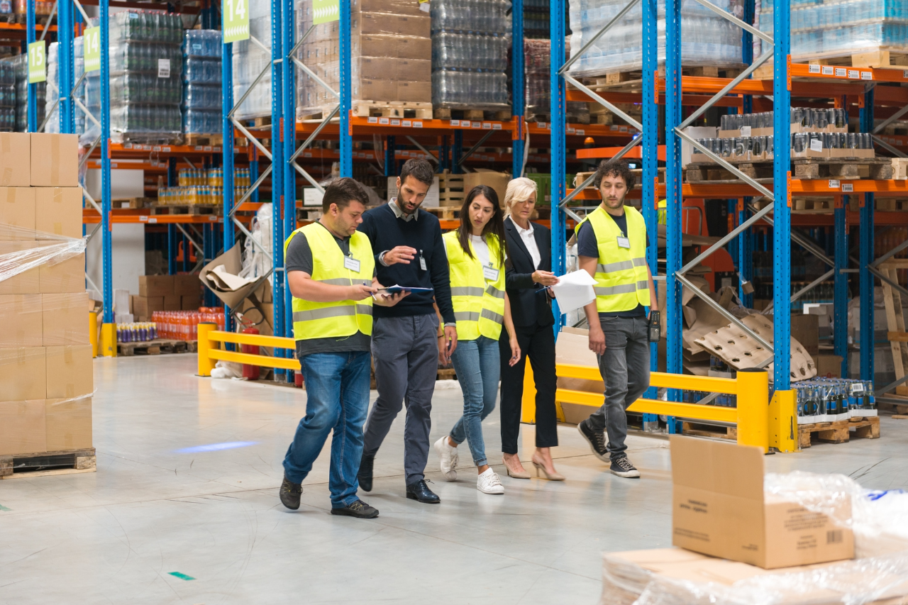 Auf dem Foto sind fünf Personen zu sehen, die in einer Reihe durch das Lager gehen. Alle sind in reflektierenden, leuchtend gelben Westen gekleidet. Die Gruppe scheint eine Inspektion oder einen Audit durchzuführen. Im Hintergrund stehen hohe Regale, auf denen Paletten mit verschiedenen Produkten und Kartons gestapelt sind.