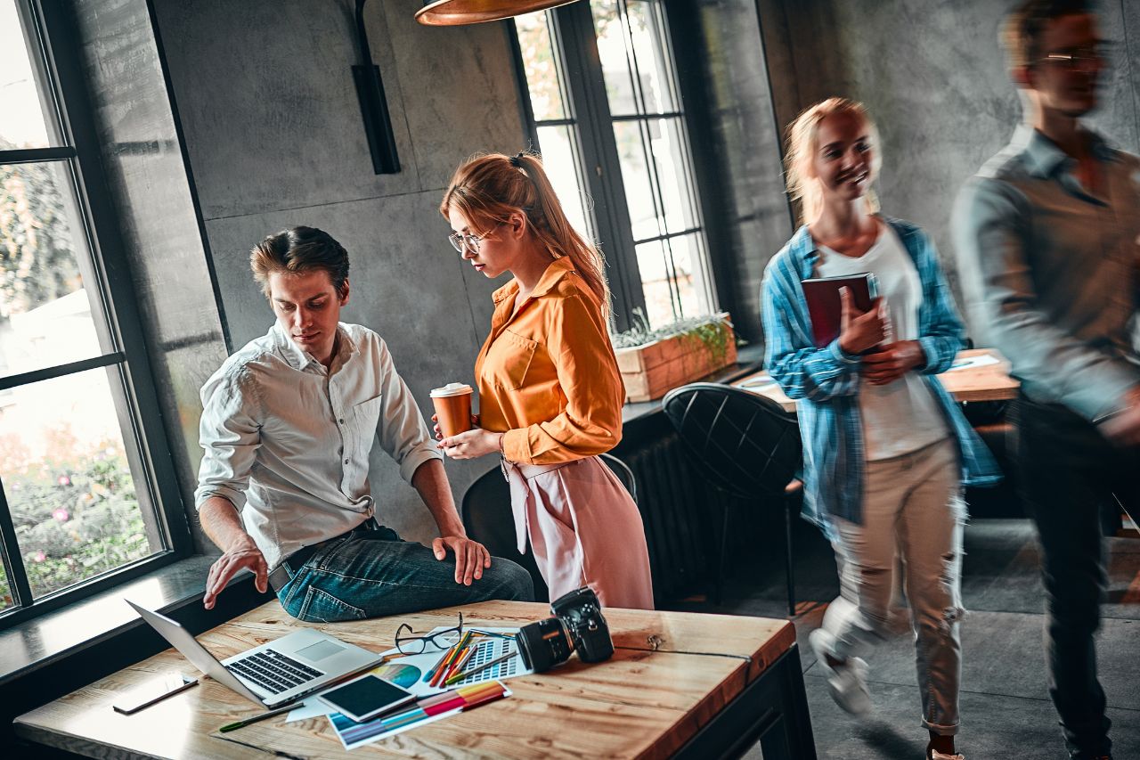 Das Bild zeigt eine Gruppe von Menschen in einem modernen, gut beleuchteten Büro oder Arbeitsbereich. Eine Person sitzt mit einem Laptop an einem Schreibtisch, eine andere Person steht neben ihr und hält eine Tasse Kaffee in der Hand. Im Hintergrund laufen zwei weitere Personen durch den Raum. Auf dem Schreibtisch liegen verschiedene Gegenstände, etwa eine Kamera, ein Notizbuch, eine Brille und farbiges Papier.