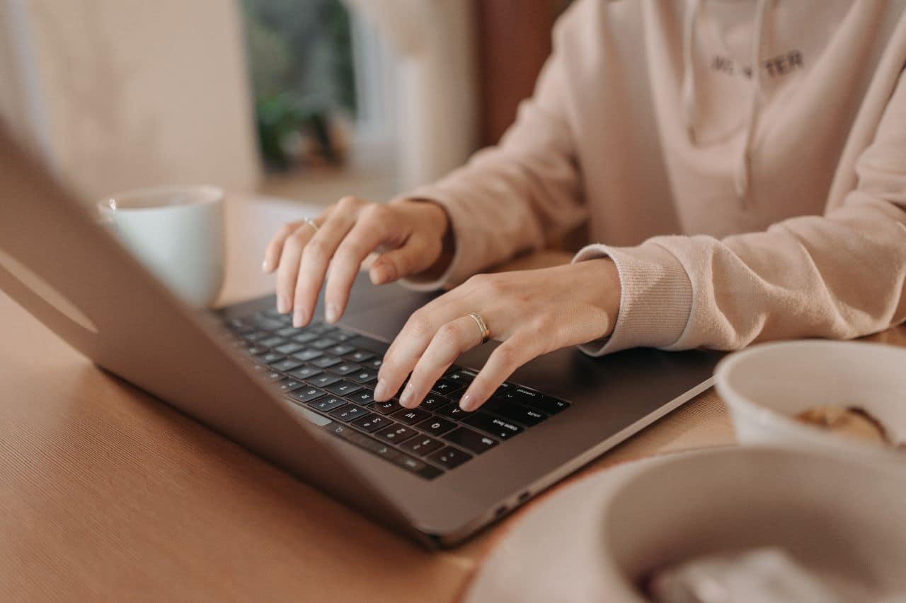 Das Bild zeigt eine Person, die auf einer Laptoptastatur tippt. Die Person trägt einen hellen Kapuzenpullover und an einem Finger ist ein Ring zu sehen. Die Szene spielt sich in einer gemütlichen Umgebung ab, möglicherweise zu Hause oder in einer anderen ungezwungenen Umgebung. Auf dem Tisch neben dem Laptop stehen eine Tasse und eine Schüssel, was die heimelige Atmosphäre unterstreicht. Der Hintergrund ist leicht unscharf, was dem Foto zusätzlich einen warmen und ruhigen Charakter verleiht.