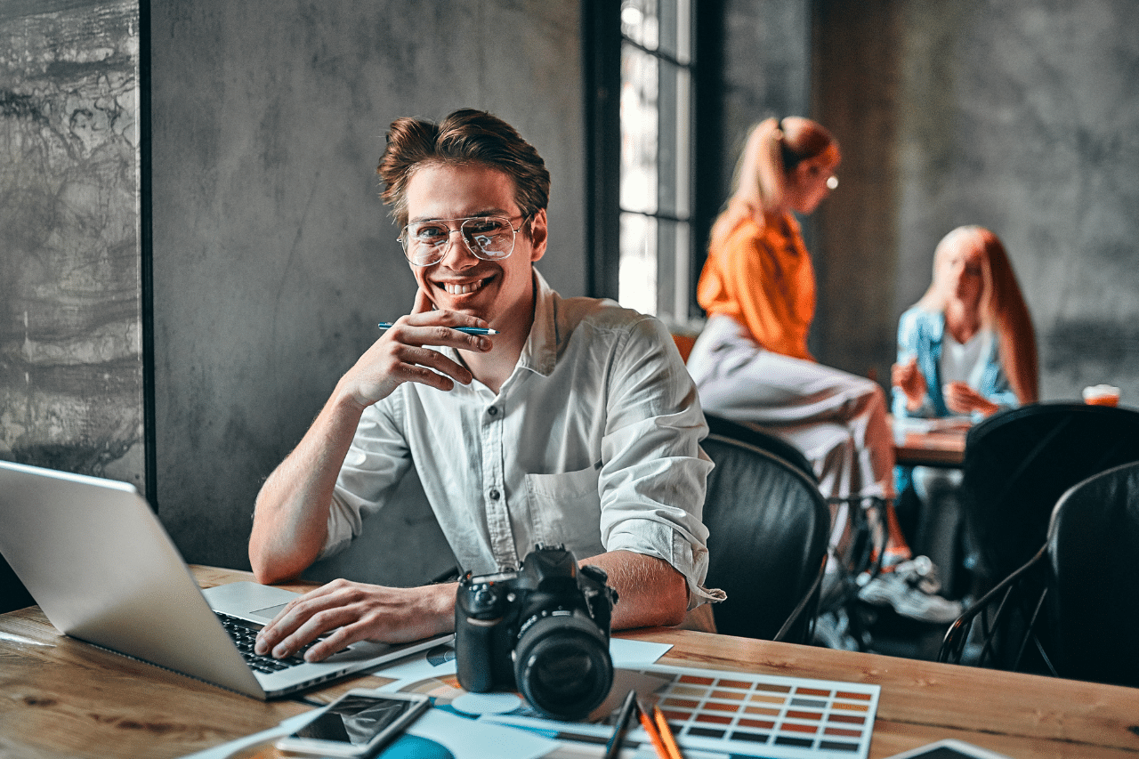 Das Gemälde zeigt einen lächelnden Mann, der an einem Holztisch sitzt und an einem Laptop arbeitet. Auf dem Tisch liegen auch eine Kamera, ein Smartphone, ein Farbwähler und einige Papiere. Im Hintergrund sind zwei weitere Personen zu sehen, die an einem anderen Tisch sitzen und sich unterhalten.
