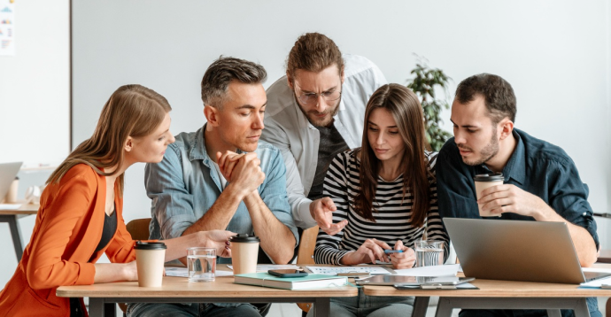 Das Bild zeigt eine Gruppe von fünf Personen, die um einen Tisch sitzen und in eine Diskussion oder Zusammenarbeit vertieft sind. Auf dem Tisch liegen mehrere Gegenstände, darunter Kaffeetassen, ein Laptop, Notizblätter und Dokumente. Eine der Personen zeigt auf etwas auf dem Tisch, was aktives Mitwirken und Zusammenarbeit andeutet.