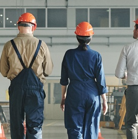 Auf dem Bild sehen wir drei Personen, die sich in einer Industrie- oder Bauumgebung von der Kamera entfernen. Zwei von ihnen tragen einen Overall und einen orangefarbenen Schutzhelm, die dritte Person trägt ein weißes Hemd und keinen Helm. In der Umgebung sehen wir Industrieanlagen und einen großen, offenen Raum, der an eine Fabrik- oder Lagerumgebung erinnert.