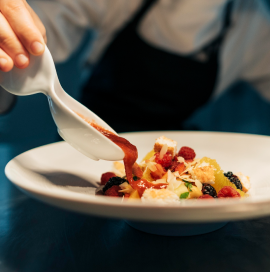 Das Foto zeigt eine Person, die Soße auf eine Schüssel auf einem weißen Teller gießt. Das Gericht wird mit einer Vielzahl von Zutaten zubereitet, darunter Früchte wie Himbeeren und Brombeeren und andere dekorative Elemente. Eine Person hält eine weiße Sauciere in der Hand, aus der rote Soße ausgegossen wird.
