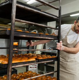 Das Foto zeigt einen Mann in einer Schürze, der in einer Bäckerei arbeitet. Der Mann bewegt ein metallenes Bücherregal mit Regalen voller frisch gebackener Brötchen. Im Hintergrund sind ein Bäckereiofen und andere Elemente der Bäckereiausrüstung zu sehen.
