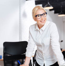 Auf dem Foto lehnt eine lächelnde Frau an einem Schreibtisch. Eine Frau mit blonden Haaren, die ein weißes Hemd und eine dunkle Hose trägt. Die Frau trägt eine Brille.