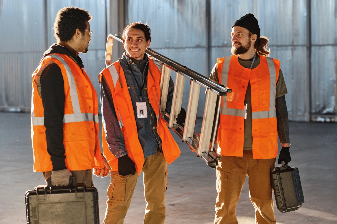 Auf dem Foto sehen wir drei Männer in reflektierenden, orangefarbenen Westen, die in einer großen, vermutlich industriellen Halle stehen. Jeder von ihnen trägt Schutzhandschuhe, und zwei von ihnen halten einen schwarzen, plastischen Werkzeugkoffer. Der Mann in der Mitte, mit einem Helm auf dem Kopf, hält eine zusammengeklappte Leiter und lächelt den Mann zu seiner rechten Seite an.