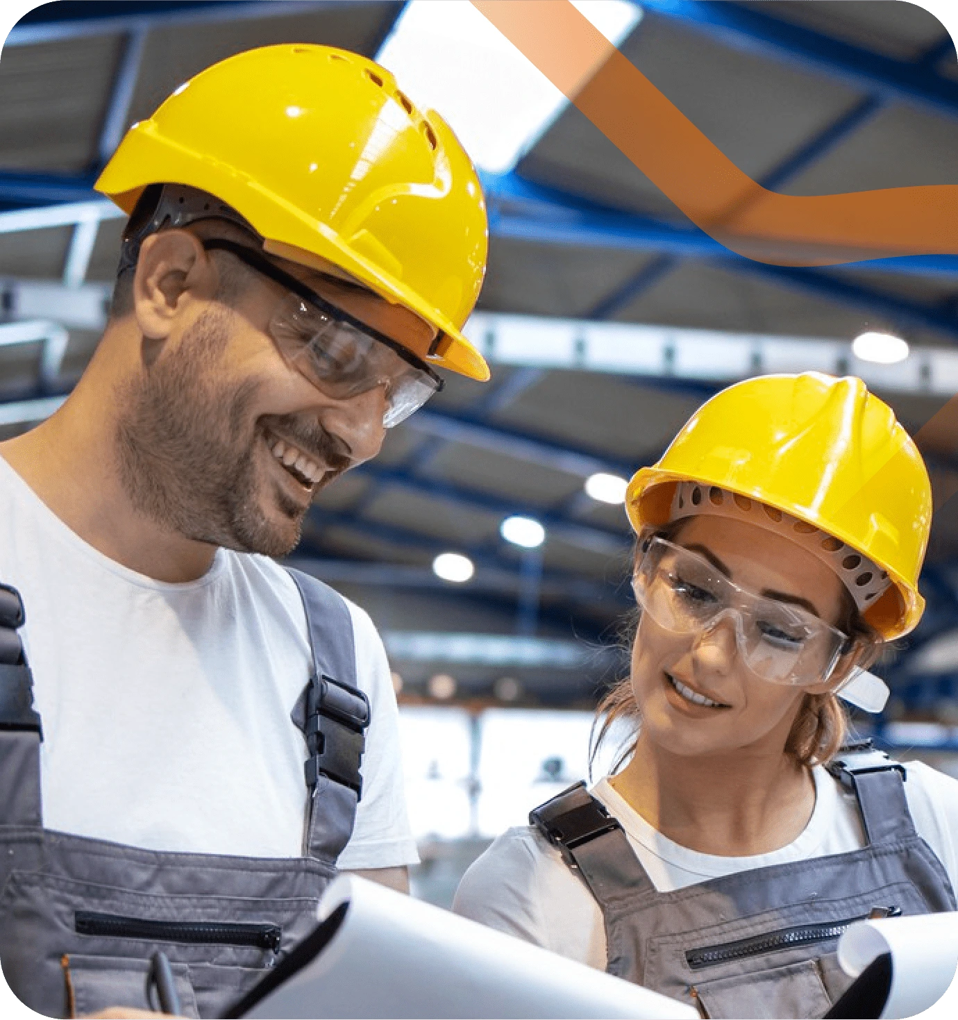 Zwei Arbeiter in Schutzhelmen und Schutzbrillen stehen in einer modernen Industriehalle. Sie tragen graue Arbeitskleidung und lächeln, während sie gemeinsam ein Dokument betrachten.