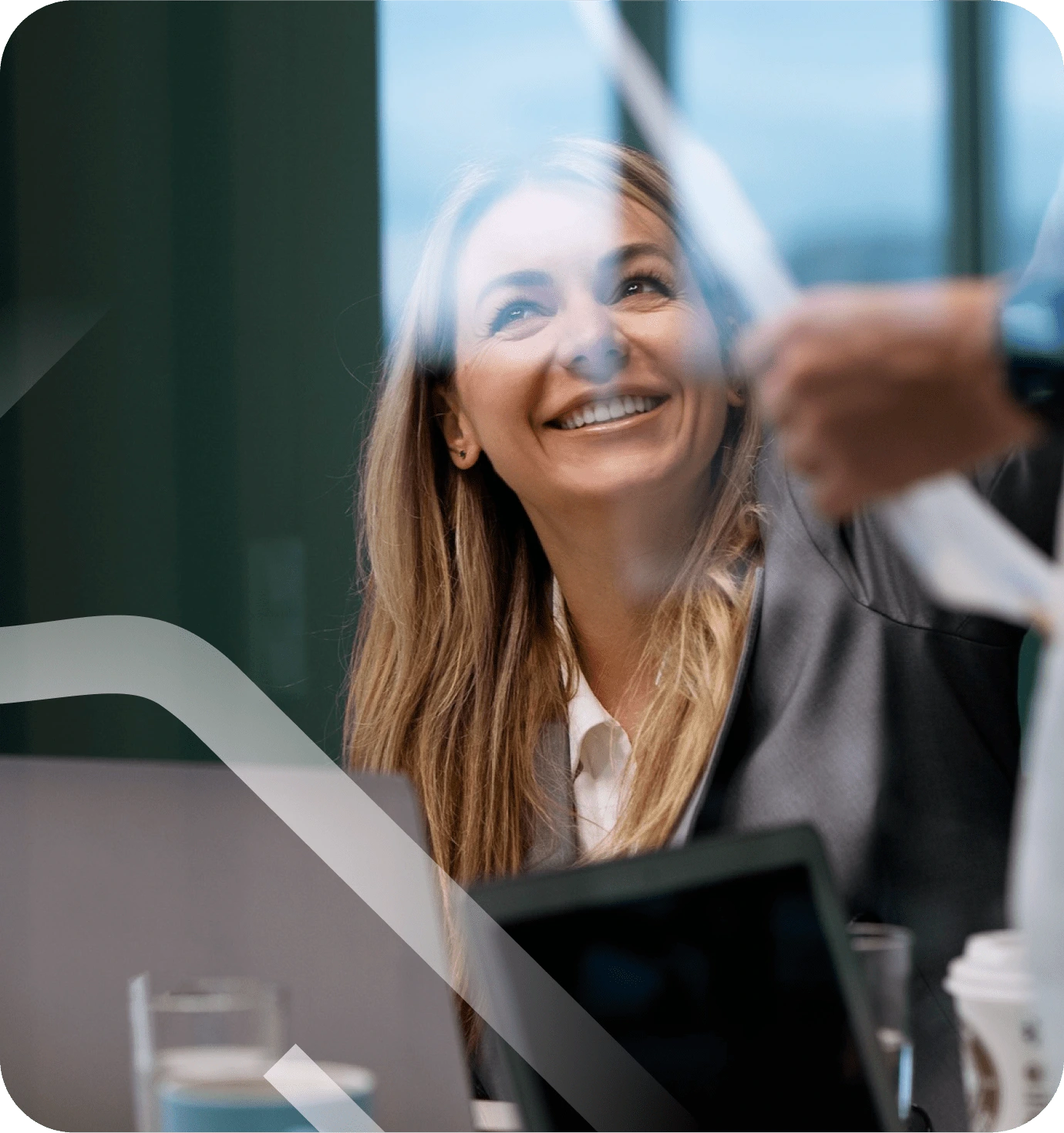 Eine lächelnde Frau mit langen, blonden Haaren sitzt in einem modernen Büro. Sie trägt einen eleganten Blazer und schaut aufmerksam auf jemanden, der ein Dokument hält. Vor ihr stehen ein Laptop und ein Glas, was auf ein Geschäftstreffen oder eine Besprechung hindeutet.