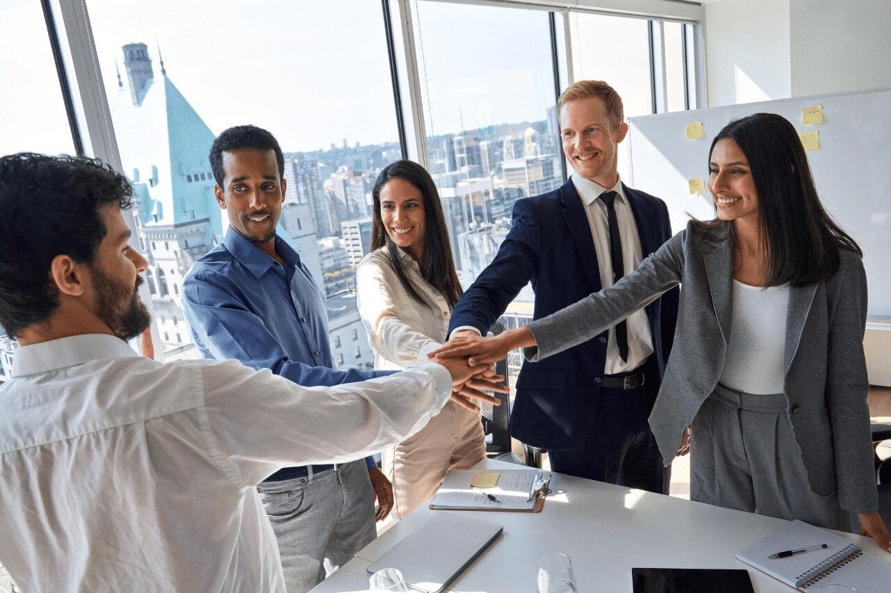 In einem modernen Büro mit großen Fenstern und Blick auf die Stadt stehen fünf beruflich gekleidete Personen um einen Tisch. Alle strecken ihre Hände zur Mitte – ein kraftvolles Symbol für Teamgeist, Zusammenhalt und gemeinsame Ziele. Die Stimmung ist positiv und engagiert, die Gesichter zeigen Lächeln und Motivation. Auf dem Tisch liegen Notizbücher, ein Tablet und Wassergläser, was auf ein produktives Meeting oder eine kreative Besprechung hindeutet. Im Hintergrund ist ein Whiteboard mit bunten Haftnotizen zu sehen – vermutlich Teil eines Brainstormings oder Projektplans. Die Szene strahlt Dynamik, Vertrauen und professionelle Zusammenarbeit aus.