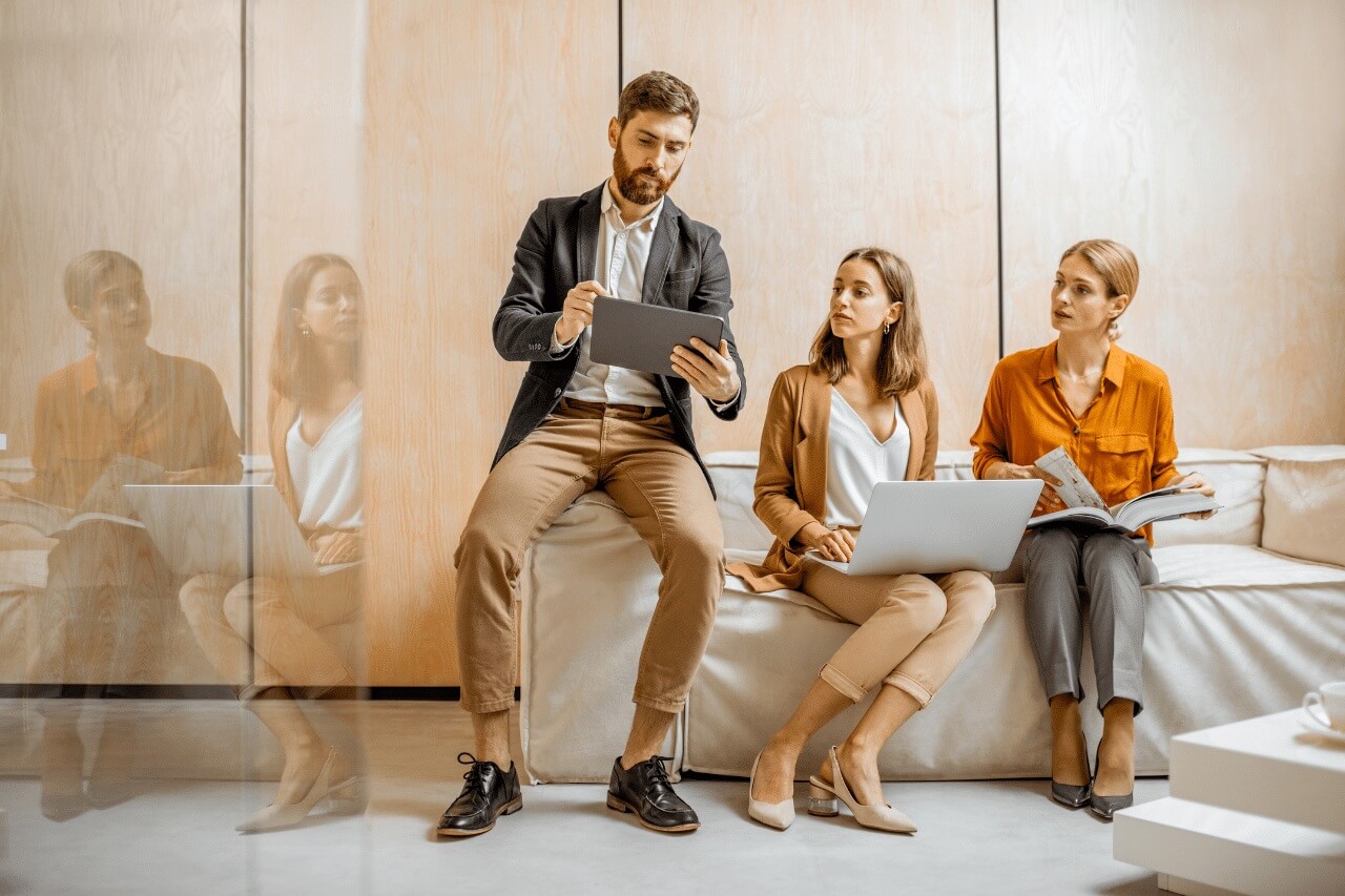 Das Foto zeigt drei Personen in einem modernen Büro. Auf der linken Seite sitzt ein Mann auf der Sofalehne, hält ein Tablet und zeigt den beiden Frauen neben ihm etwas. Er trägt eine dunkle Jacke, ein weißes Hemd und beigefarbene Hosen. Die Frau in der Mitte hat einen Laptop auf dem Schoß und hört aufmerksam zu. Sie trägt ein weißes Top und eine hellbraune Jacke sowie passende Hosen. Auf der rechten Seite sitzt eine andere Frau mit einem geöffneten Buch oder Notizbuch. Sie trägt eine orangefarbene Bluse und graue Hosen. Es scheint, dass alle drei an einem Treffen oder einer Diskussion teilnehmen. Der Raum ist hell, modern und minimalistisch, mit Holzpaneelen an den Wänden und einem hellen Sofa.