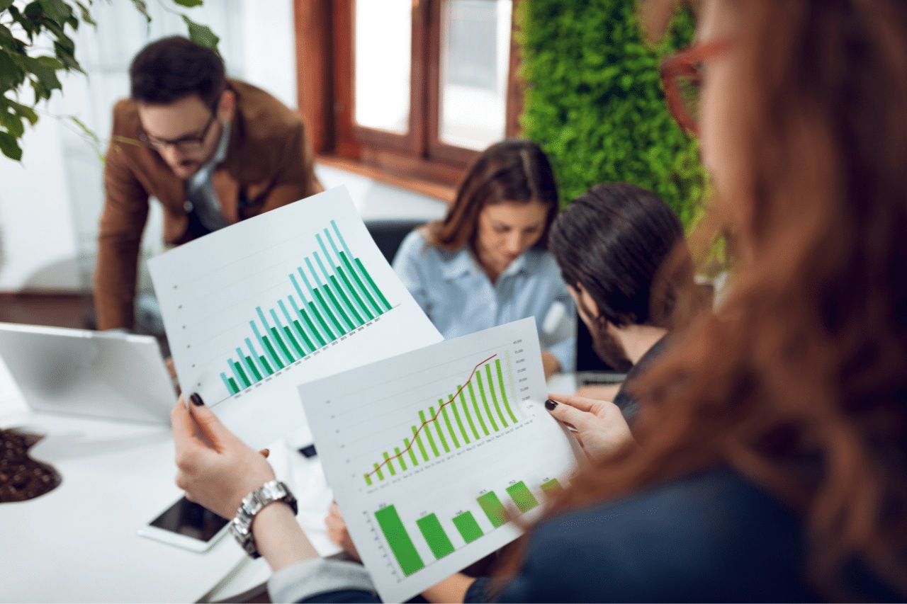 W nowoczesnym, jasnym biurze cztery osoby pracują razem nad projektem. Na pierwszym planie jedna osoba trzyma dwie kartki papieru z wykresami słupkowymi i liniowymi, prezentującymi zielone i niebieskie serie danych w czasie – wyraźny dowód analizy opartej na danych. Pozostali trzej członkowie zespołu siedzą wokół stołu z laptopami i dokumentami, pogrążeni w dyskusji. Środowisko jest otwarte i przyjazne: rośliny doniczkowe i zielona ściana wprowadzają do pomieszczenia naturalne elementy i tworzą inspirującą, zrównoważoną atmosferę pracy. Scena ta odzwierciedla skupienie, współpracę i wykorzystanie nowoczesnych technologii w codziennym życiu biurowym.