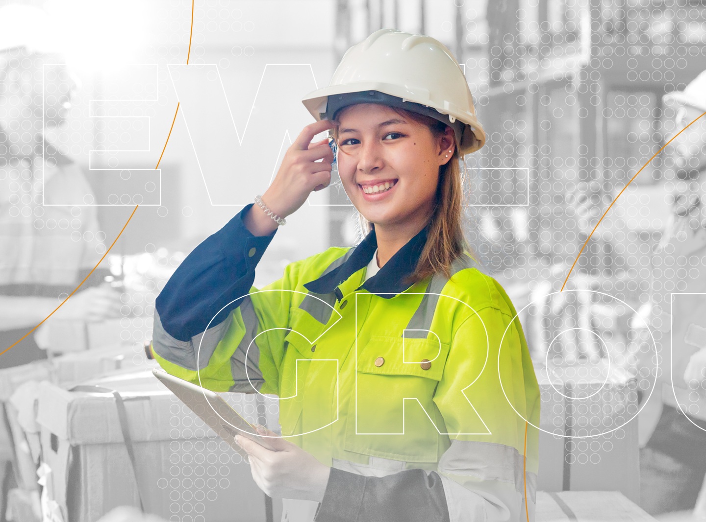 Die lächelnde Frau trägt einen weißen Schutzhelm und eine auffällige Arbeitsjacke in Gelb und Dunkelblau – typische Kleidung für Sicherheitsbereiche wie Bauwesen, Logistik oder Industrie. In der Hand hält sie ein Klemmbrett, bereit für Kontrolle, Koordination oder Dokumentation. Im Hintergrund sind weitere Personen und Arbeitsmaterialien wie Kisten oder Container zu sehen, was auf eine dynamische, teamorientierte Umgebung hinweist.