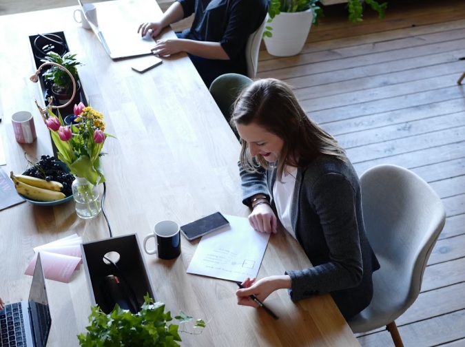 In einem warmen, einladenden Arbeitsraum sitzen zwei Personen an einem langen Holztisch. Die Atmosphäre wirkt entspannt und kreativ – eine Mischung aus Konzentration und Leichtigkeit. Im Vordergrund schreibt jemand auf ein Blatt Papier, auf dem ein klar strukturierter Ablaufplan zu sehen ist: „Start“, „Idea“, „Research“, „Plan“, „Execute“ – ein typischer Workflow für Projektentwicklung oder kreative Prozesse. Im Hintergrund arbeitet eine zweite Person am Laptop. Der Tisch ist liebevoll dekoriert: eine Vase mit Blumen, eine Schale mit Obst, eine Kaffeetasse, ein Notizbuch und eine kleine Topfpflanze schaffen eine wohnliche, fast inspirierende Umgebung. Natürliches Licht fällt auf den Holzfußboden und verstärkt die freundliche Stimmung.