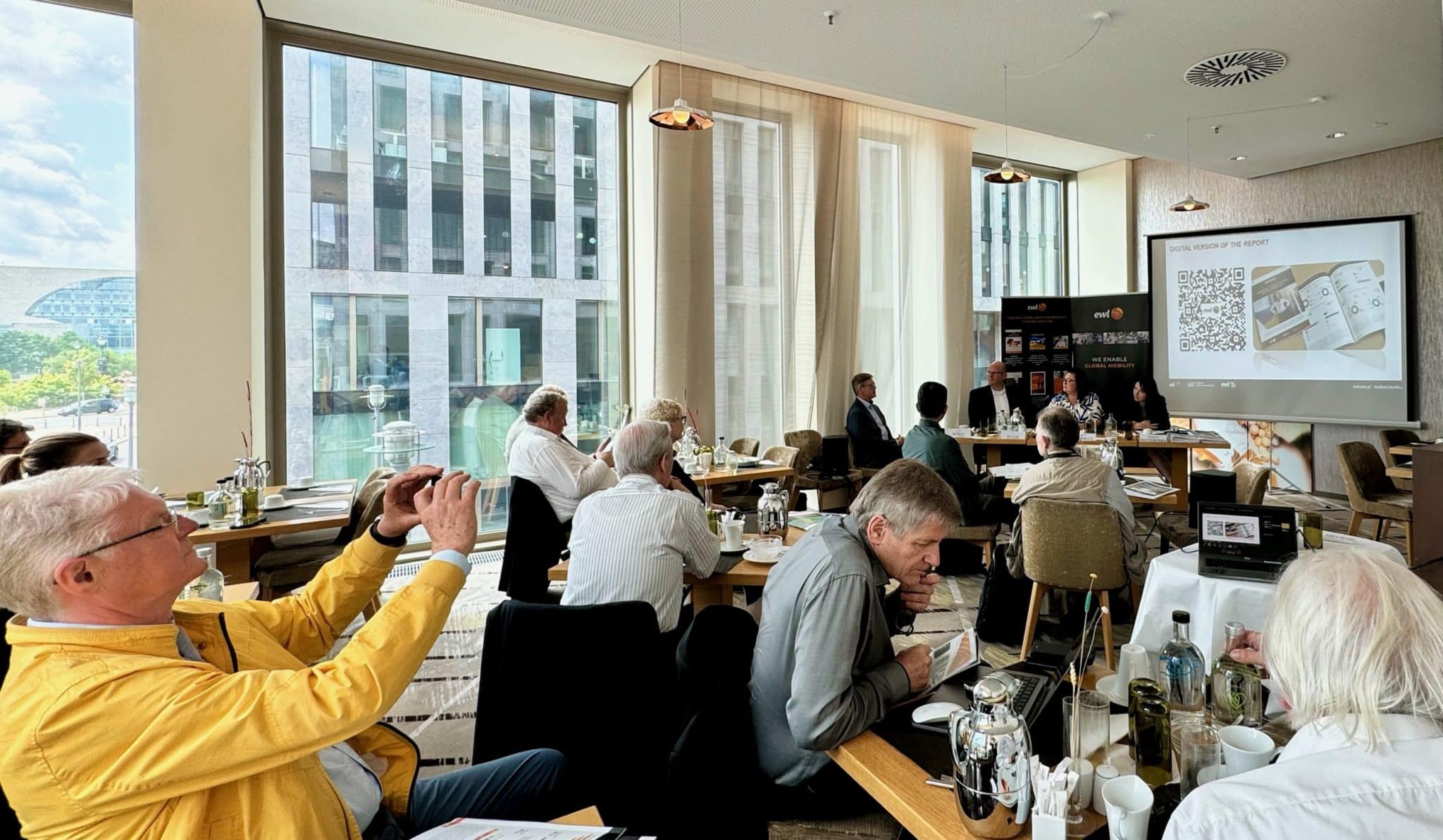 W nowoczesnej, jasnej sali konferencyjnej z dużymi oknami odbywa się seminarium lub panel dyskusyjny. Kilku uczestników siedzi przy stołach, uważnie słuchając panelu z przodu sali. Osoba w żółtej kurtce trzyma smartfon, dokumentując moment – za pomocą zdjęcia lub filmu. Na ekranie wyświetlany jest slajd prezentacji z kodem QR i tytułem „Przyszłość sztucznej inteligencji w Europie”. Butelki z wodą, szklanki i notatniki na stołach podkreślają profesjonalną atmosferę. Scena ta oddaje poczucie skupienia, wymiany wiedzy i postępu technologicznego w kontekście europejskim.