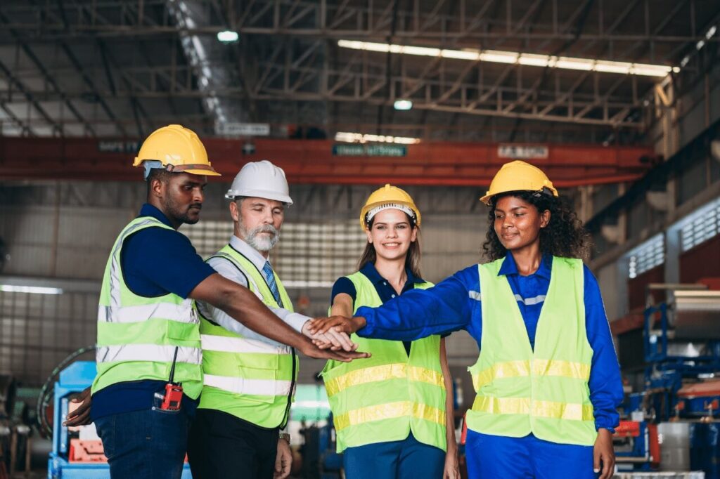 Das Foto zeigt vier lächelnde Personen, die in einer großen Produktions- oder Industriehalle stehen. Alle tragen gelbe Sicherheitswesten und Helme – drei gelbe und einen weißen. Die Person mit dem weißen Helm und Hemd scheint eine Vorgesetzte oder für technische Angelegenheiten verantwortliche Person zu sein. In einer Geste der Einheit legen alle ihre Hände übereinander – ein Symbol für Zusammenhalt, Vertrauen und gemeinsame Verantwortung.