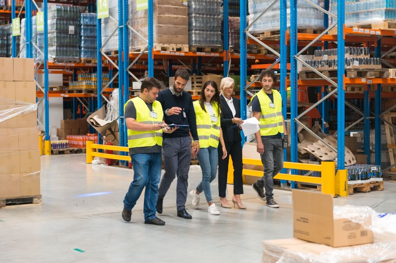 Fünf Personen gehen nebeneinander durch ein geräumiges, gut organisiertes Lagerhaus mit hohen Regalen aus blauen und orangefarbenen Metallstreben. Die Regale sind sorgfältig mit Kartons und Flaschen bestückt – ein klarer Hinweis auf strukturierte Logistik und effizientes Bestandsmanagement. Drei der Personen tragen gelbe Warnwesten und wirken wie Mitarbeitende oder Sicherheitsverantwortliche. Die anderen zwei sind in Businesskleidung gekleidet und halten Dokumente sowie ein Tablet – vermutlich Führungskräfte oder externe Besucher bei einer Inspektion oder einem Rundgang.
