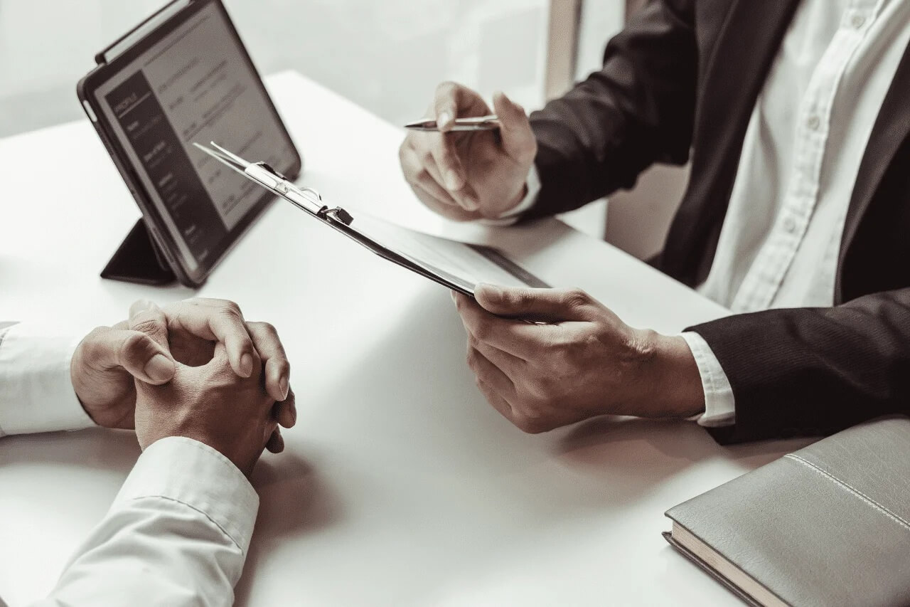 Zwei Personen sitzen sich an einem weißen Tisch gegenüber – die Szene wirkt wie ein Bewerbungsgespräch oder ein strategisches Meeting. Die Person im dunklen Anzug hält ein Klemmbrett und einen Stift, offenbar in der Rolle des Gesprächsführers oder Evaluators. Gegenüber sitzt jemand in weißem Hemd, mit gefalteten Händen, aufmerksam und konzentriert. Auf dem Tisch liegen ein Tablet mit sichtbarem Text, ein geschlossenes Notizbuch und das Klemmbrett mit Unterlagen.