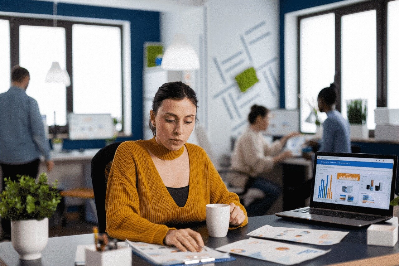 Das Foto zeigt ein modernes Büro, in dem mehrere Personen konzentriert arbeiten. Im Vordergrund sitzt eine Frau in einem senffarbenen Pullover an einem Schreibtisch, hält eine weiße Tasse in der Hand und analysiert ausgedruckte Diagramme. Auf dem Bildschirm eines Laptops ist ein Dashboard mit dem Titel „Mitarbeiter-Management-Dashboard“ und farbigen Balkendiagrammen zu sehen. Der Raum ist hell, lichtdurchflutet und mit Pflanzen und geometrischen Ornamenten dekoriert.